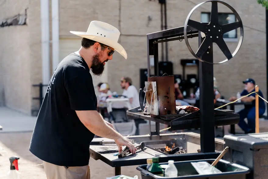 A pitmaster in a straw hat working an offset smoker